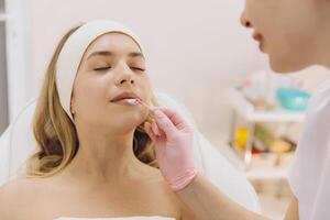 Beautician carefully applying anesthetic cream on client's lips using wooden stick, preparing for permanent makeup procedure in beauty salon photo