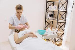 Beautician wearing pink gloves applying cosmetic product on young woman face lying on massage table in beauty salon photo