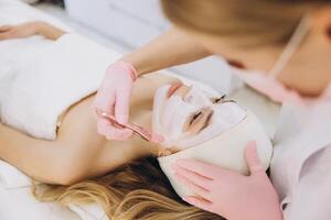 Beautician in gloves applying a facial mask to a client in a spa setting, focusing on skincare and relaxation. The client is lying down, wrapped in a towel photo