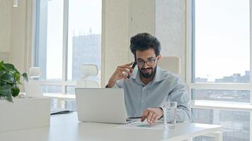 Busy multitasking Arabian businessman manager consultant Indian muslim man talk mobile phone check information on laptop computer work consulting client smartphone call business consultation in office photo