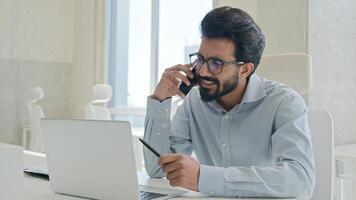 Busy multitasking Arabian businessman manager consultant Indian muslim man talk mobile phone check information on laptop computer work consulting client smartphone call business consultation in office photo