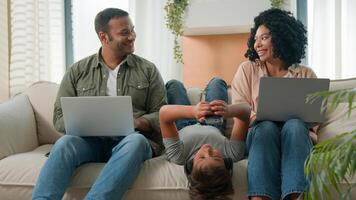 Addicted family use gadgets devices little kid child son boy lying upside down on sofa play game on mobile phone smiling African American parents working laptops different gen using modern tech photo
