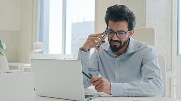 Busy multitasking Arabian businessman manager consultant Indian muslim man talk mobile phone check information on laptop computer work consulting client smartphone call business consultation in office photo