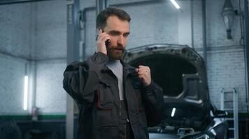 Annoyed serious Caucasian adult 30s man in uniform auto mechanic engineer talking mobile phone in repair car service male worker of technical industry communicate speak smartphone at vehicle workshop photo