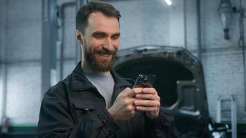 Smiling cheerful man 30s Caucasian guy auto mechanic technical factory industry worker engineer in uniform using mobile phone looking at smartphone screen laughing work repair service at car garage photo