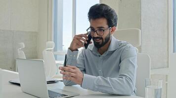 Busy multitasking Arabian businessman manager consultant Indian muslim man talk mobile phone check information on laptop computer work consulting client smartphone call business consultation in office photo
