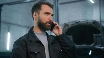 Caucasian man male guy mechanic in car service station industrial worker in uniform talking phone smartphone conversation answer mobile call talk with client explain automobile problem order details photo