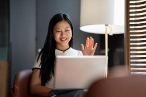 A smiling Asian woman has an online meeting with her team while working remotely at a coffee shop. photo