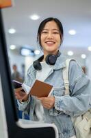 A smiling Asian female passenger stands in front of an airport self-check-in kiosk in the airport. photo
