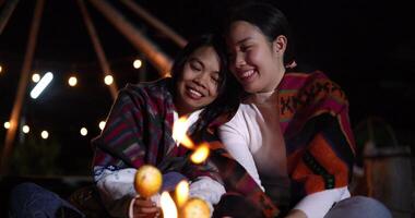 Handheld selective focus shot, Two Asian siblings enjoy talking and grilling corn while sitting around a warm campfire on a chilly evening video
