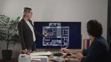 A woman is giving a presentation of an architectural project. On the large screen, there is a house plan with furniture layout. A male colleague is listening to the designer and commenting video