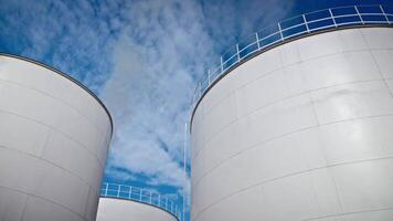 Large industrial storage tanks under a bright blue sky with scattered clouds. The white cylindrical structures feature metal railings on top, standing out against the vibrant backdrop. video