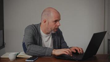 A middle-aged man is at home working on his laptop. He is typing intensively and looking at the monitor with focus and concentration, fully immersed in his work. video