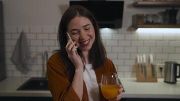 Beautiful brunette woman in a modern kitchen is talking on the phone and smiling. In front of her on the table are a glass of orange juice, some fruits, and a blender. video