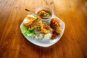 A delicious Indonesian meal featuring white rice, fried chicken, fried tempeh, lettuce, crackers, and a bowl of vegetable soup on a wooden table photo