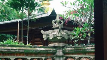 A traditional Balinese architectural feature, adorned with intricate carvings, surrounded by lush greenery and tropical plants. The background shows a wooden structure typical of Balinese design video