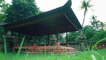 A traditional Balinese pavilion surrounded by lush greenery, featuring intricate stone carvings and a thatched roof. The pavilion is set in a serene garden environment video