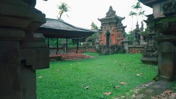 A serene Balinese temple courtyard featuring traditional architecture, lush green grass, and scattered leaves. The scene includes ornate stone structures and a pavilion video