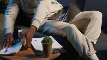 Casual person sitting On the sofa and reading research papers with analytics, multitasking with laptop at home. Man diving into professional or personal topics of improvement, independent. Camera A. video