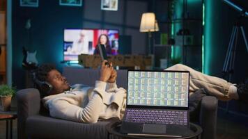 Man enjoying music and reading e books on tablet while waiting for stock market financial figures to get updated on laptop. Person at home next to pc showing trading investment graphs. Camera B. video