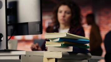 Focus on stack of books on startup office desk with woman in blurred background typing on PC, solving tasks. Pile of books in venture company workspace in front of employee working, camera B closeup video