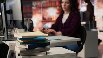 Focus on stack of books on startup office desk with woman in blurred background typing on PC, solving tasks. Pile of books in venture company workspace in front of employee working, camera A closeup video