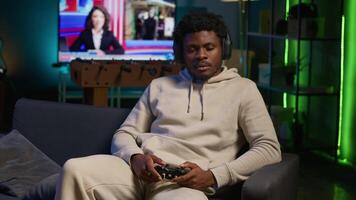 Bored guy plays online game and spamming attack buttons on his controller, modern loft apartment. Gamer in neon lit space participating in internet e sports competition. Camera A. video