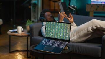 African american person using his tablet laying on the couch while he pays attention to laptop with stock market data trends. Focused man comparing trading graphs numbers before investing. Camera A. video
