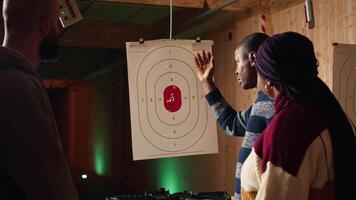 Shooting gallery instructor explaining bullseye scoring system to customers. African american firing range specialist teaching clients bullseye points system, discussing industry regulations, camera A video