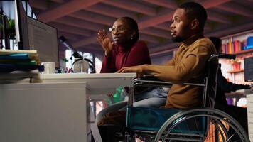 Manager coming to inclusive startup office employee in wheelchair desk to offer help. Thoughtful supervisor checking up on software development agency worker with disability, camera B video