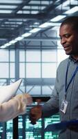 Vertical Data center engineer greeting colleague, examining system diagnostics paperwork together. Server room workers doing handshake, meeting to look over maintenance reports, camera B video