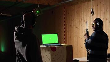 Green screen laptop next to shooting gallery safety officer watching client shoot at targets, supervising him. Mockup notebook and firing range expert teaching man how to use firearms, camera A video