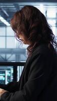 Vertical Admin in data center using laptop to update storage arrays, configuring network virtualization. Woman in server room holding coffee cup, verifying settings to maintain stable performance, camera B video