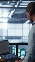 Vertical Engineer in data center operating notebook, deploying new server architectures. Server farm employee verifying settings using laptop, maintaining stable network performance, camera B video