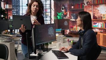 Startup intern holding laptop asks questions to senior technician coding advanced AI algorithm. Employee requests help from CTO manager in Silicon Valley workplace, camera A video