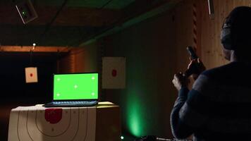 Green screen laptop next to man in firing range pressing mechanism to recall paper targets. Mockup notebook and person training with handguns on bullseye, sharpening skills, camera A video