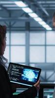 Vertical Data center technicians deploying AI driven management tools using tablet. Server farm coworkers inspecting server clusters, updating network topologies using artificial intelligence, camera A video