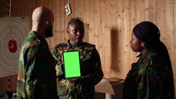 Soldiers at shooting range do training designed to increase safety and survivability in combat, watching instructional videos on green screen chroma key tablet device presented by commander, camera A