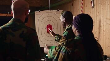 Army commander teaching recruits in shooting gallery how to shoot paper targets, explaining pointing system. Military officer instructing soldiers on how to hit bullseye, reading rules, camera A video
