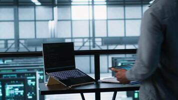 Admin arriving in data center, using artificial intelligence tech on tablet and laptop. Man in server farm using AI on devices to update storage arrays, improving performance, camera A video