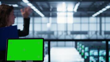 Focus on chroma key device in front of data center technicians analyzing metrics. Close up of isolated screen tablet next to server hub workers ensuring seamless operations, camera B video