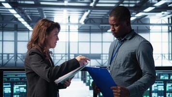 Data center team examines paperwork files in server room, reviewing system diagnostics. Server room workers looking over maintenance schedules and cybersecurity protocols, camera B video
