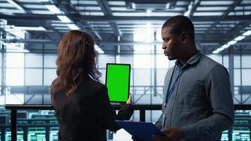 Computer scientists in data center use mockup tablet, inspecting server rigs, updating devices. Teamworking IT colleagues collaborating to ensure stable operations, using chroma key device, camera B video