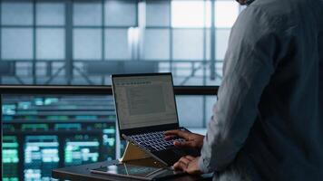 Engineer in data center operating notebook, deploying new server architectures. Server farm employee verifying settings using laptop, maintaining stable network performance, camera A video