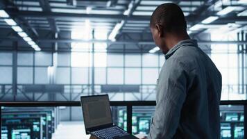 Engineer in data center operating notebook, deploying new server architectures. Server farm employee verifying settings using laptop, maintaining stable network performance, camera B video