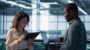 Smiling admins using laptop in data center, managing server virtualization. Cheerful IT specialist and african american man using notebook in server room, ensuring infrastructure reliability, camera A video