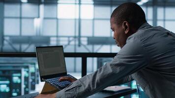 Technician coding on laptop in data center, managing server virtualization, finetuning network protocols. Server farm employee programming on notebook, enforcing advanced security measures, camera A video