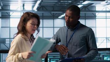 Team of IT professionals looking over data center checkup checklist paperwork. African american man and colleague collaborating in server farm reading documentation files, camera A video