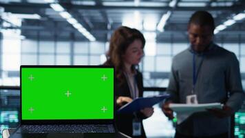 Green screen laptop used by data center admins in blurry background reviewing equipment diagnostics. Chroma key notebook used by coworkers adjusting server room configurations, camera B video