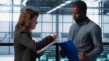 Data center team examines paperwork files in server room, reviewing system diagnostics. Server room workers looking over maintenance schedules and cybersecurity protocols, camera A video